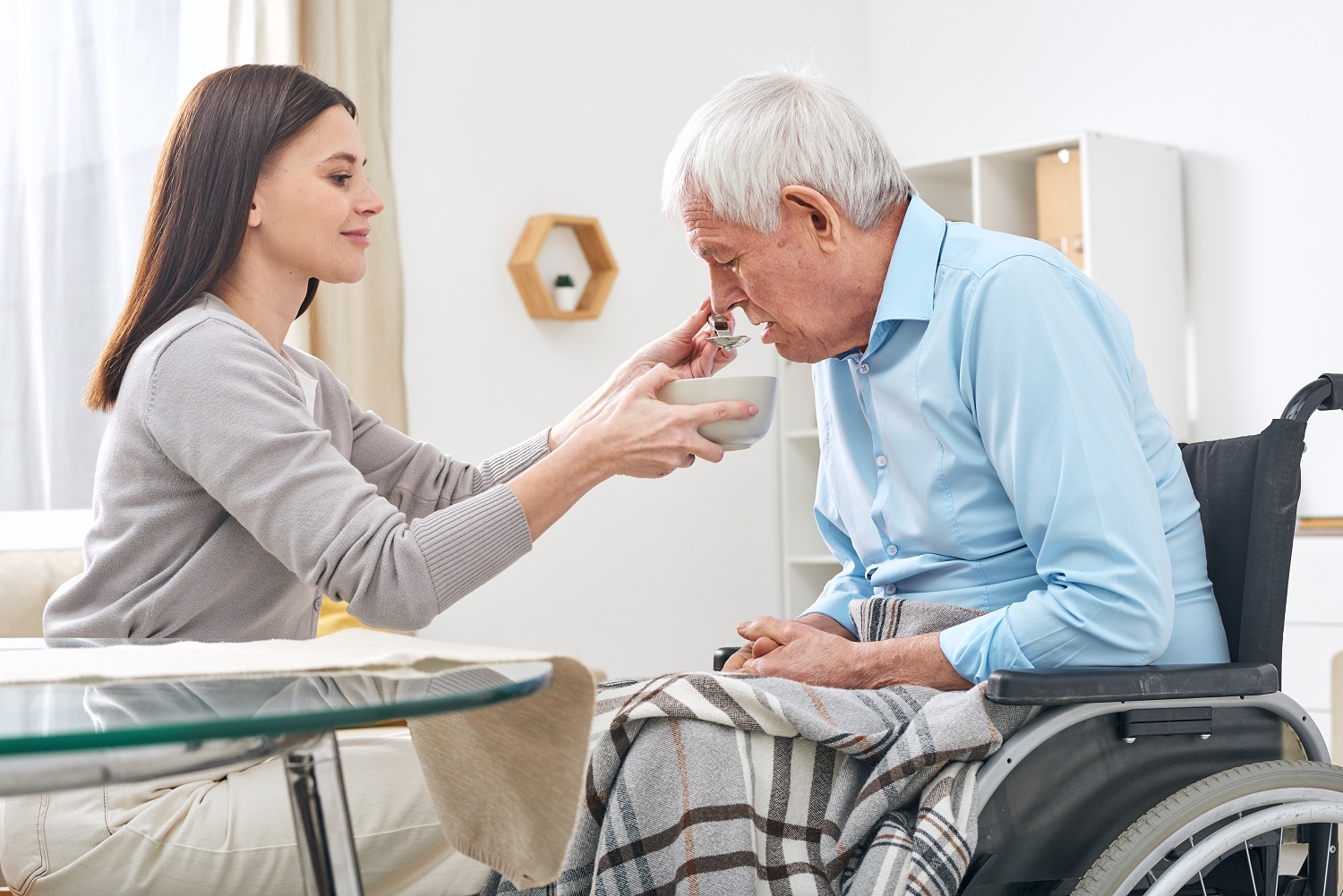 Social worker holding bowl with soup and spoon by mouth of senior disable man